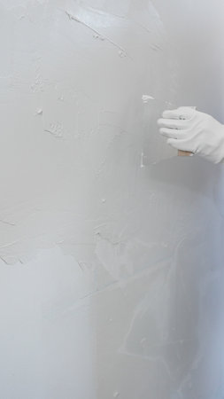 Construction worker wearing gloves and blue overalls is using a putty knife to apply plaster on a white wall, performing home renovation and improvement, close-up of handの写真素材