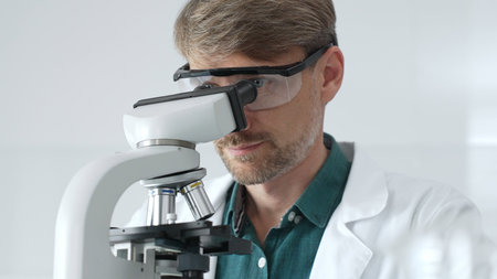 Senior man scientist wearing protective glasses and lab coat analyzing samples with a microscope in...