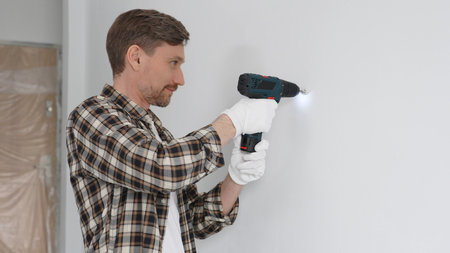 Male construction worker in checked shirt and safety gloves is screwing a screw into the wall using a cordless screwdriver, carrying out a careful home renovation. Portrait viewの写真素材