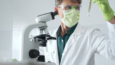 Masked senior man researcher examining green botanical samples in test tube in laboratory during...