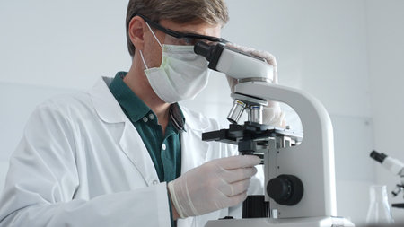 Adult male scientist wearing white mask, lab coat and protective glasses using microscope, doing...