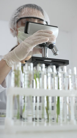 Female scientist wearing white protective gear using microscope, analyzing test tubes in laboratory, showing scientific research and development. Medicine and science conceptの写真素材