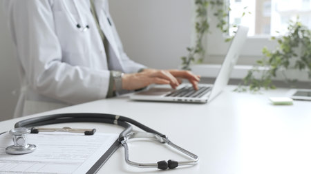 Stethoscope and medical records are on the desk opposite the doctor is typing on a laptop with a in...