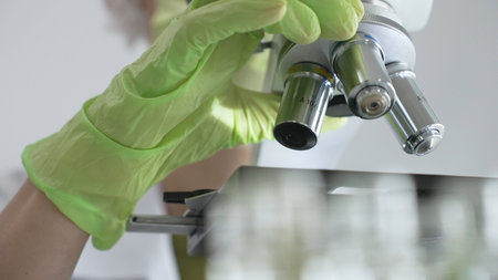 Close up of green gloved hands of a scientist adjusting a microscope in a laboratory, emphasizing...