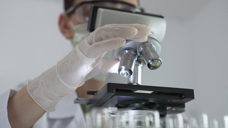 Female scientist wearing white protective gear analyzing test samples under microscope in...