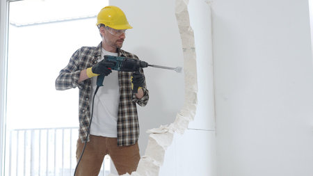 Male construction worker wearing beige checkered shirt, yellow hard hat and protective gloves, is demolishing white wall with rotary hammer drill, generating dust, medium long shot. Renovation projectの写真素材