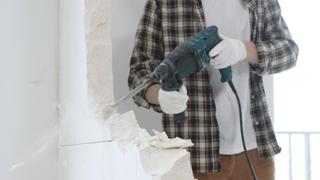 Close up of unrecognizable male construction worker wearing beige checkered shirt and protective gloves, is demolishing white wall with rotary hammer drill, generating dust, close up view. Renovationの写真素材