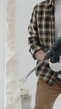 Close up of unrecognizable male construction worker wearing beige checkered shirt and protective gloves, is demolishing white wall with rotary hammer drill, generating dust. Renovation conceptの写真素材