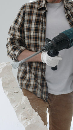 Close up of unknown male construction worker wearing beige checkered shirt and protective gloves, is demolishing white wall with rotary hammer drill, generating dust. Renovation projectの写真素材