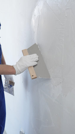 Close up of the construction worker hand wearing protective gloves, spreading plaster smoothly across wall using professional drywall taping knife realizing renovation project, vertical viewの写真素材