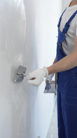 Professional construction worker wearing blue work overall is using a finishing trowel to carefully...