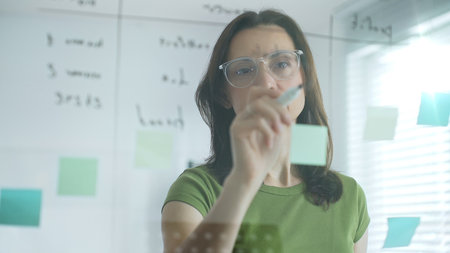 Businesswoman writing notes on glass board, surrounded by sticky papers during strategic planning...