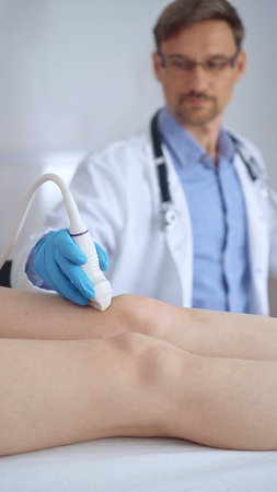 Doctor man with blue protective gloves using ultrasound equipment on the knee of a female patient,...