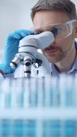 Man scientist with protective glasses and gloves is using microscope in laboratory, vertical...