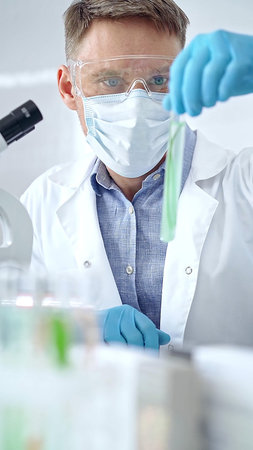 Scientist with protective gloves and glasses examining lab tube with a green fluid. Microbiology and...