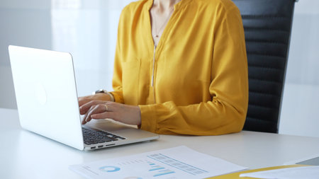 Unknown businesswoman wearing yellow blouse working on laptop at minimalist white desk in contemporary workplace, typing with focus. Business people conceptの写真素材