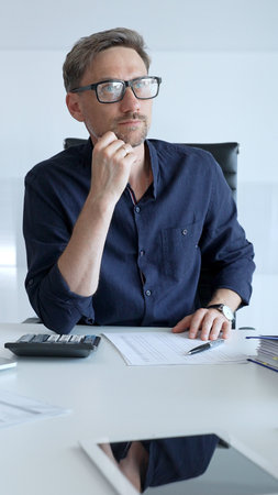 Businessman sitting at a sleek desk, working on a laptop and thinking in a bright, modern office filled with natural light. Business people conceptの写真素材