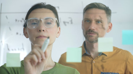 Professional woman with glasses marking glass board, arranging sticky notes while male colleague...