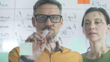 Professional team members brainstorming strategy, writing notes on transparent whiteboard with...
