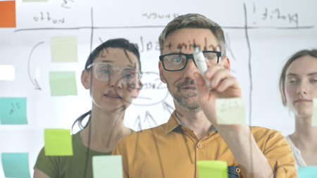 Business colleagues brainstorming, writing strategy notes on glass board, collaborating with...