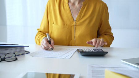 Female accountant wearing yellow shirt is sitting at white desk and using calculator while taking...