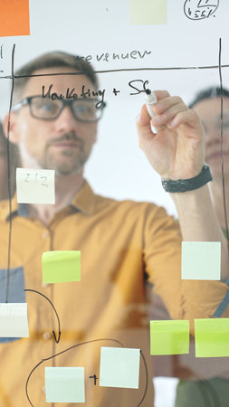 Business team collaborating on a project, using a glass board with sticky notes and marker to...
