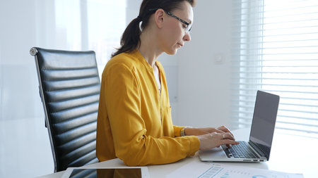 Professional businesswoman wearing yellow shirt and eyeglasses working intently, typing on laptop in...