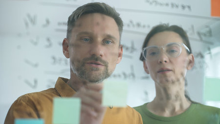 Business colleagues collaborating, using sticky notes on glass board, brainstorming innovative...