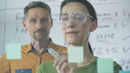 Businesswoman wearing glasses, writing on a glass board and organizing sticky notes, collaborates...