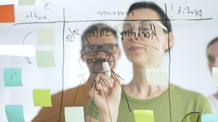 Professional team brainstorming, sharing strategy on glass whiteboard with colorful sticky notes,...