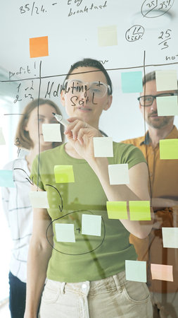 Professional businesswoman wearing glasses writing on glass board, marking strategic plan with...