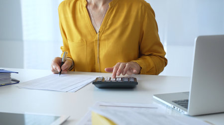 Female accountant wearing yellow shirt is sitting at white desk and using calculator while taking...
