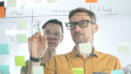 Business colleagues brainstorming, writing strategy notes on glass board, collaborating with...