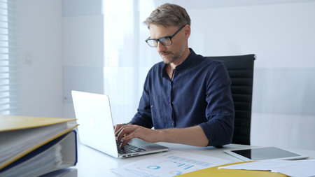 Professional businessman wearing glasses, intensely working on laptop, typing amid modern office...