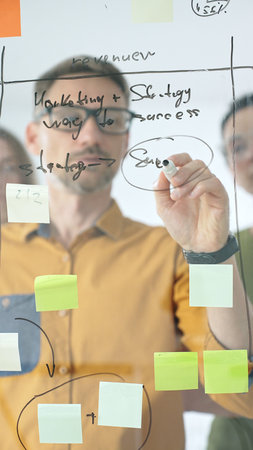 Business colleagues planning strategy, writing notes on glass wall, collaborating with enthusiasm in...