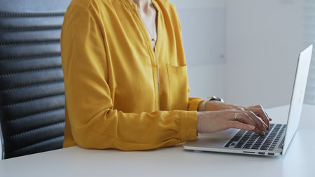 Businesswoman wearing yellow shirt sitting at white desk typing on laptop in bright modern office, working productively on computer. Business people conceptの写真素材