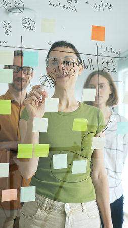 Professional businesswoman wearing glasses writing on glass board, marking strategic plan with...