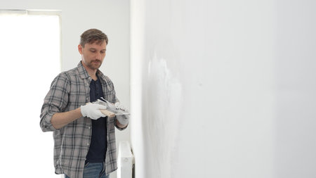 Male construction worker wearing protective gloves and plaid shirt, spreading plaster smoothly...
