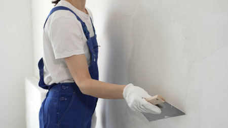 Unknown construction worker woman in blue overalls smoothing wall surface, applying plaster with putty knife, wearing protective white gloves during home renovation workの写真素材