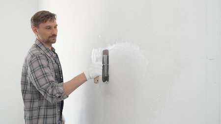Middle aged man construction worker wearing gloves and plaid shirt is using finishing trowel to plaster white wall, applying a layer of plaster to create a smooth surface during renovation projectの写真素材