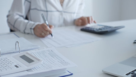 White calculator is on financial documents on the office desk, with an accountant taking notes, emphasizing accounting, finance, and tax preparation, close up. Audit and taxes conceptの写真素材