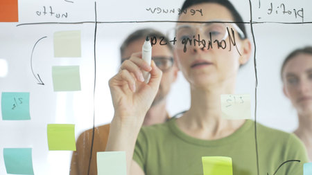 Professional businesswoman wearing glasses writing on glass board, marking strategic plan with...