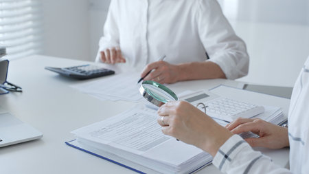 Two accountants are working together, using a magnifying glass and a calculator, to audit financial reports in their brightly lit office. Audit and taxes in businessの写真素材