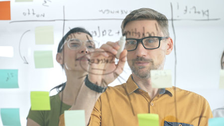 Professional team brainstorming project strategy, placing colorful sticky notes on glass board, analyzing data, sharing insights, developing collaborative solutionsの写真素材