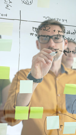 Business colleagues planning strategy, writing notes on glass wall, collaborating with enthusiasm in...