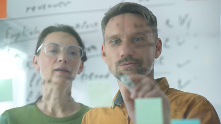 Business colleagues collaborating, using sticky notes on glass board, brainstorming innovative...
