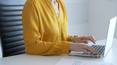 Unknown businesswoman wearing yellow blouse working on laptop at minimalist white desk in...