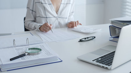 Female accountant is working with financial documents, using calculator and taking notes, with a magnifying glass and a folder on a white table in a bright office. Audit and taxes in businessの写真素材