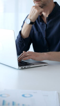 Businessman sitting at a sleek desk, working on a laptop and thinking in a bright, modern office...