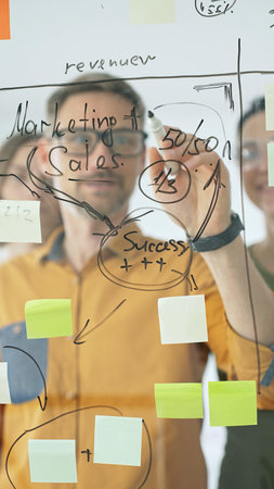 Business colleagues planning strategy, writing notes on glass wall, collaborating with enthusiasm in bright contemporary workspaceの写真素材